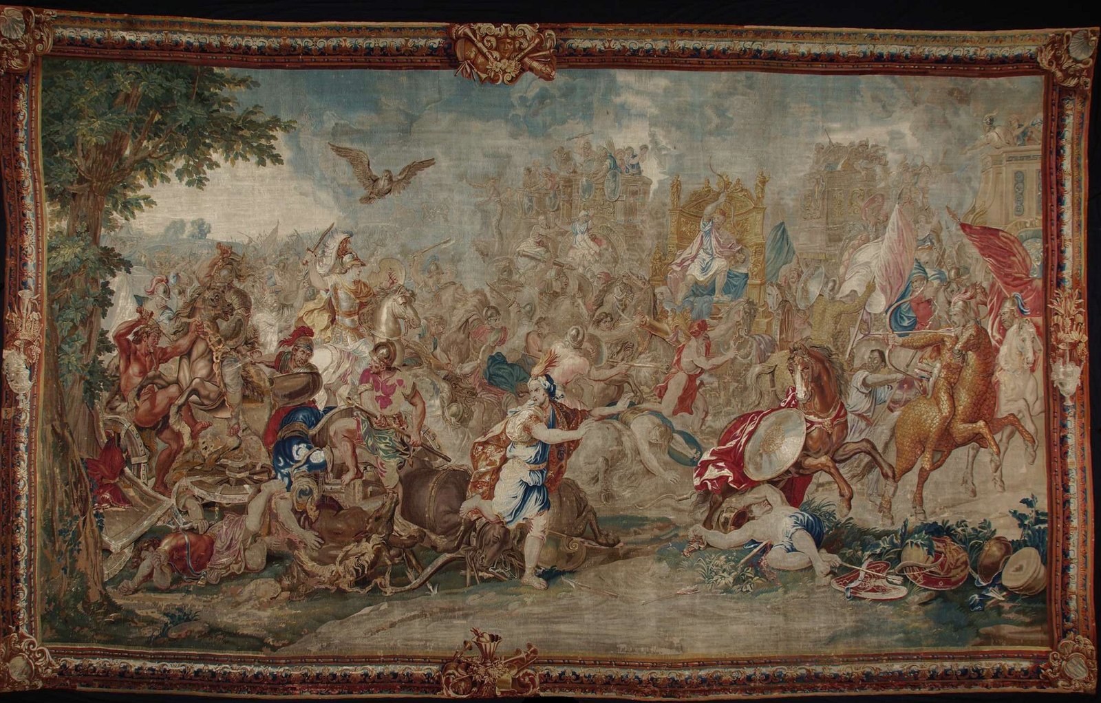 Woven tapestry in a richly decorated border: Alexander in a plumed helmet rides into a crowded mass of horsemen, chariots and fallen soldiers, while Darius in a tall golden chariot at the center turns to flee and an eagle flies overhead.