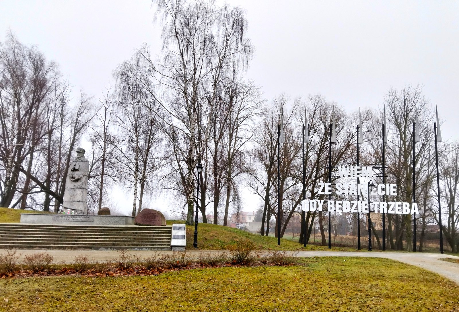 A gray stone statue of General Kleeberg in a peaked cap stands on a granite plinth in a winter park in Kock, beside bare birch trees, flagpoles and large white letters reading “Wiem, że staniecie, gdy będzie trzeba”, under an overcast sky.