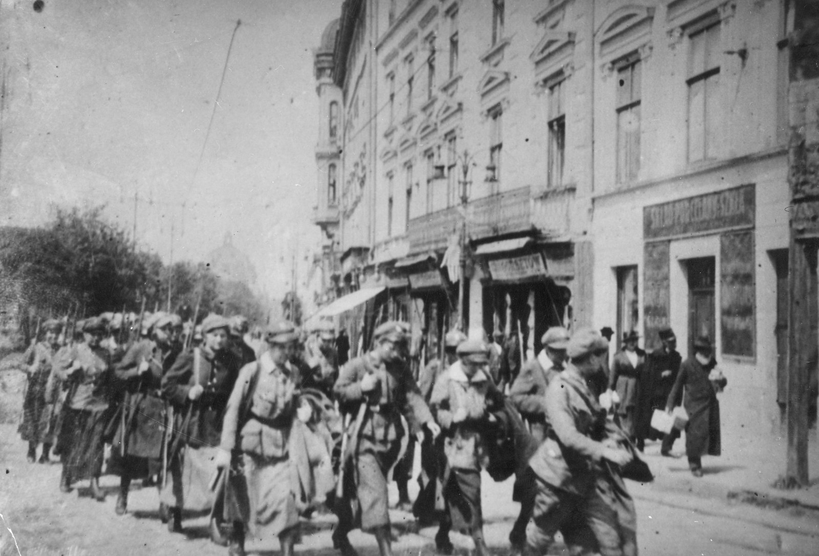 Czarno-białe zdjęcie: kolumna młodych kobiet w wojskowych czapkach i długich płaszczach, z karabinami na ramionach, maszeruje ulicą miasta wzdłuż wysokich zdobionych kamienic i szyldów sklepów, a z chodnika przyglądają się cywile.