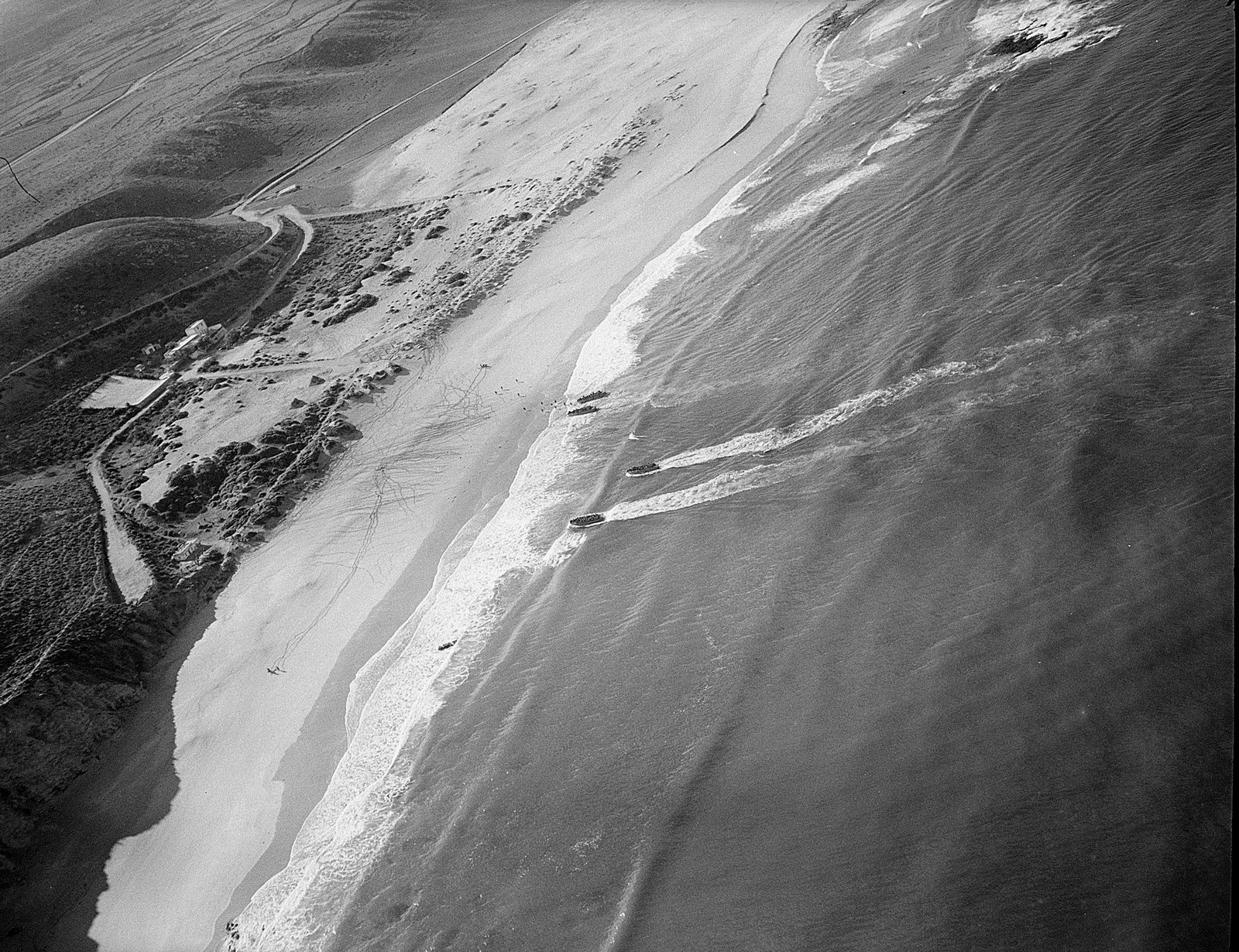 Black and white aerial photograph of a long sandy beach backed by dunes and scrubby hills: small landing craft trail white wakes through the surf toward the shore, a few already beached, with a track and a cluster of buildings inland.