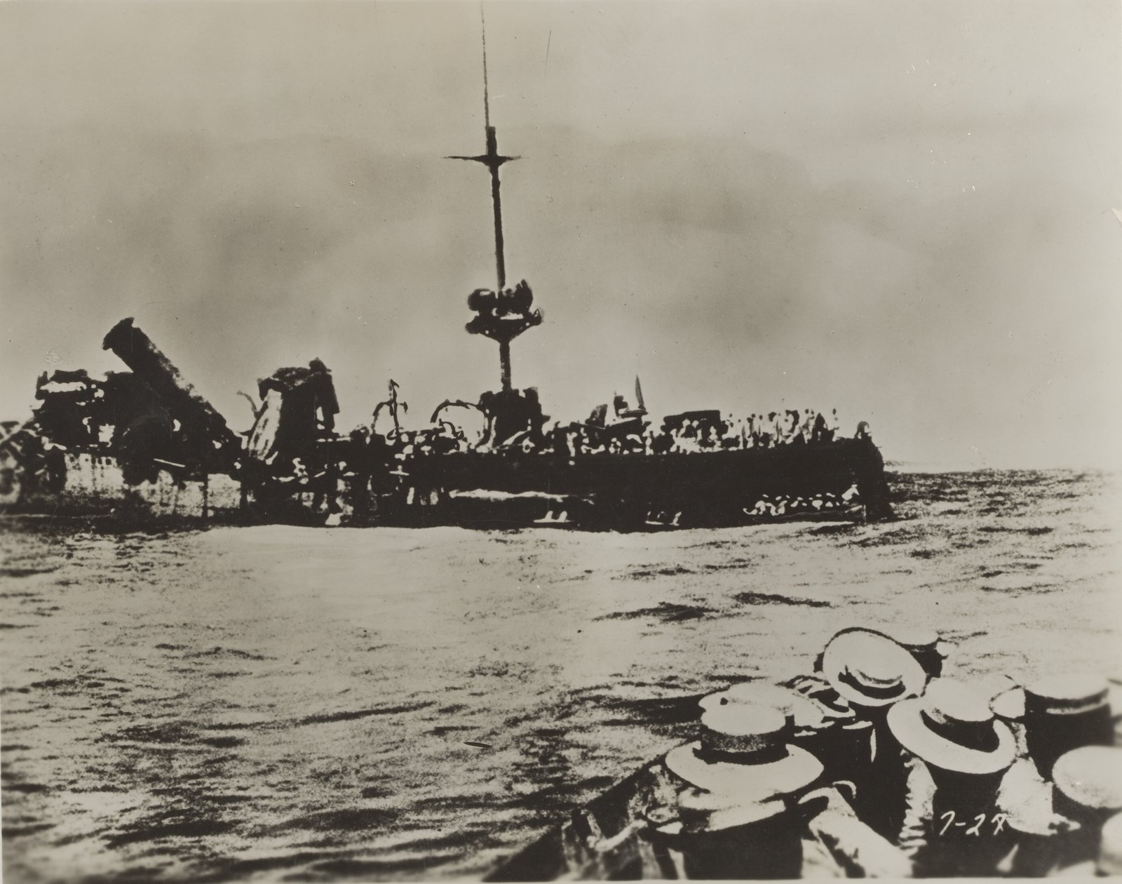 Black and white photograph taken from a boat full of sailors in straw hats, seen from behind in the foreground, approaching the shattered wreck of a warship lying low in the sea, its funnels gone, its deck a tangle of twisted metal and a single mast still standing.