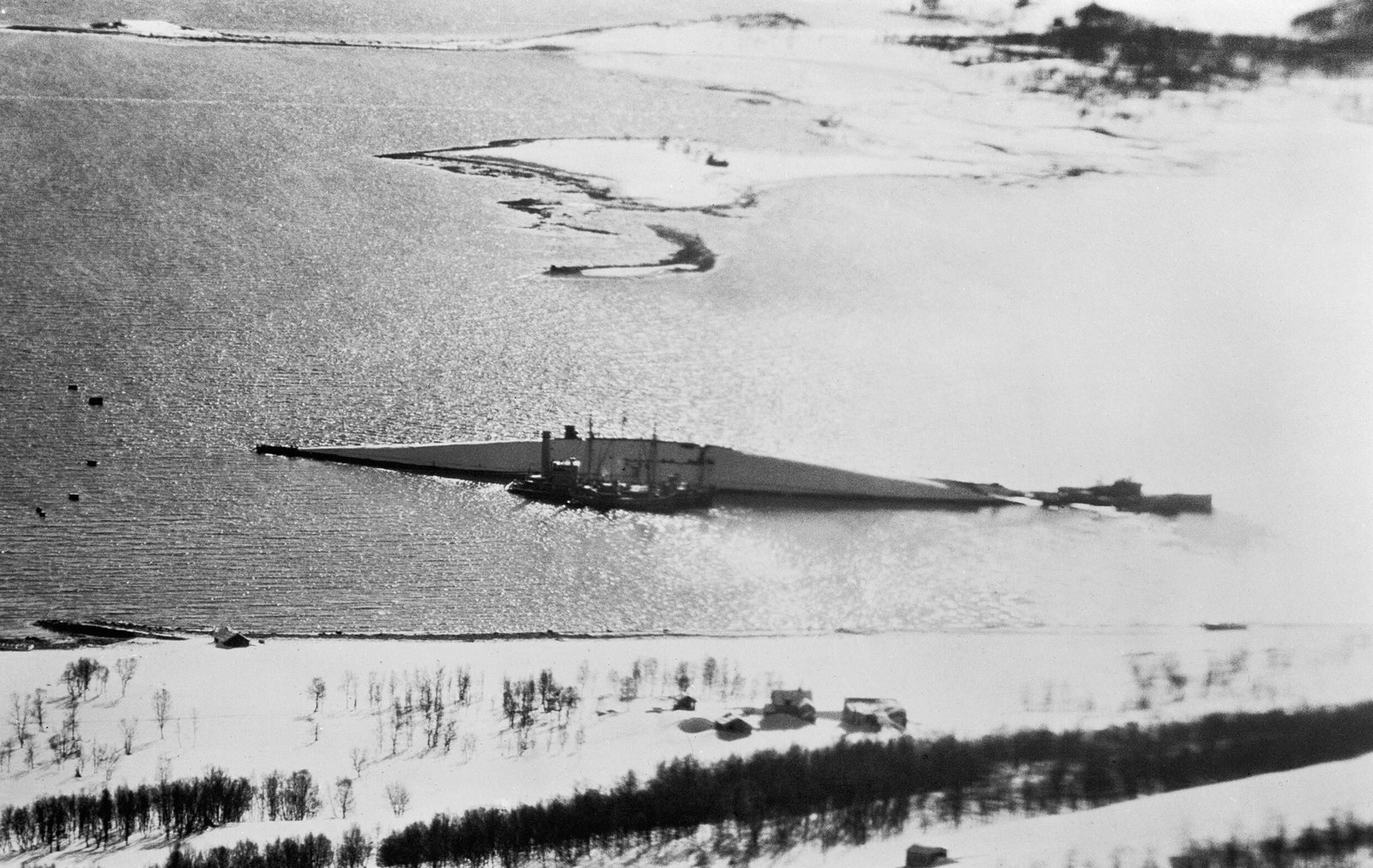 Black and white aerial photograph of a snowy fjord shore: the long upturned hull of a capsized battleship lies in shallow water glittering in low sunlight, with small salvage vessels moored alongside, and snow-covered fields and bare birch trees in the foreground.