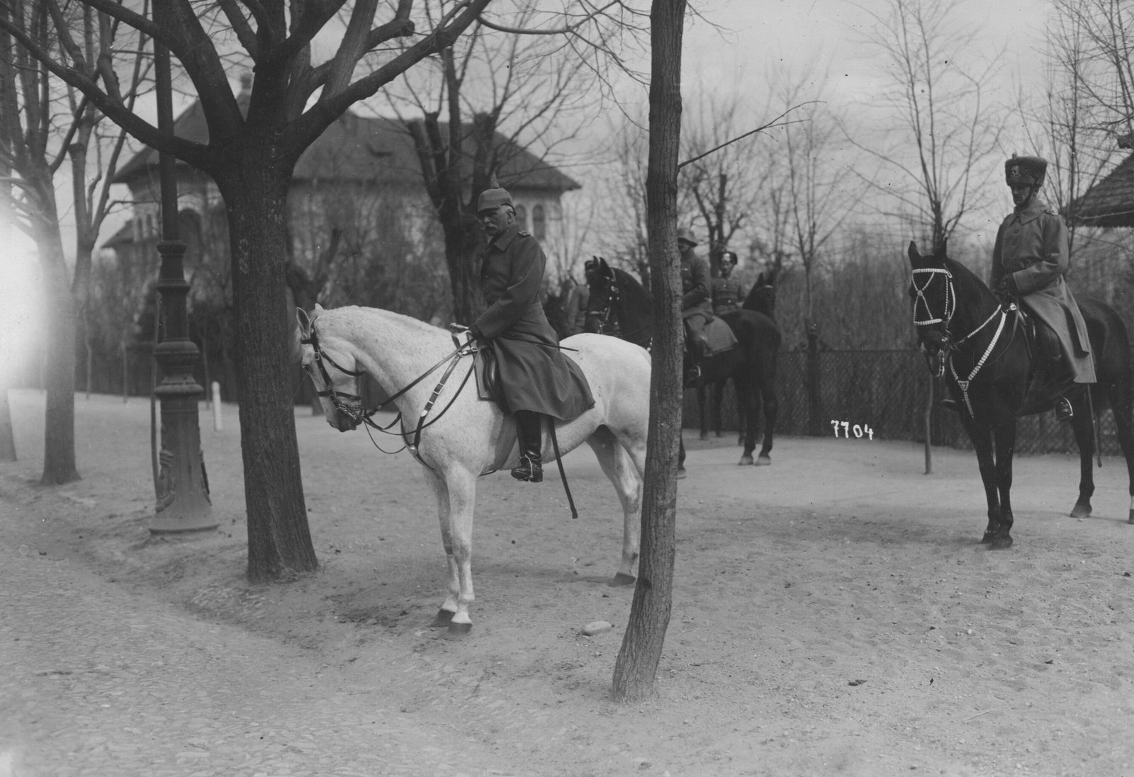 Black and white photograph of an elderly German officer in a spiked helmet and long greatcoat sitting on a white horse under bare winter trees on a sandy avenue, with mounted officers in fur caps and helmets waiting behind him and a villa in the background.