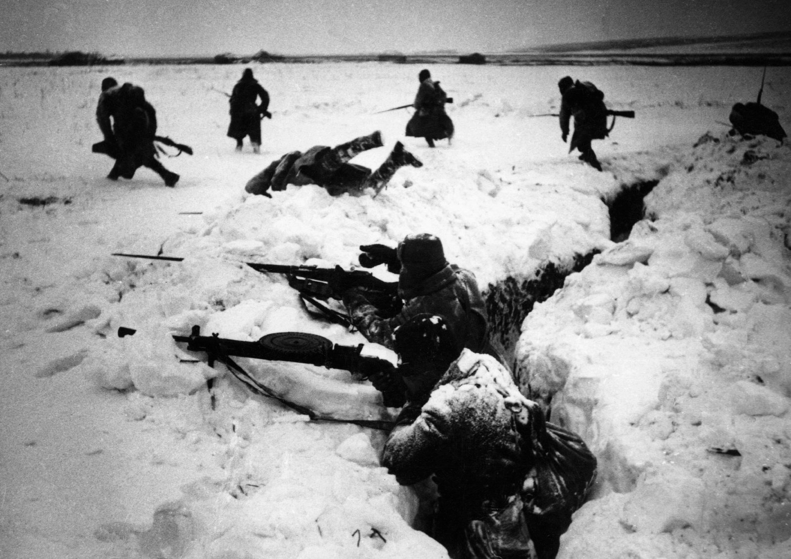 Black and white photograph of Soviet soldiers in winter coats and fur caps climbing out of a snow trench and running forward with rifles across a flat white field, while two men with a rifle and a drum-fed machine gun cover them from the trench.
