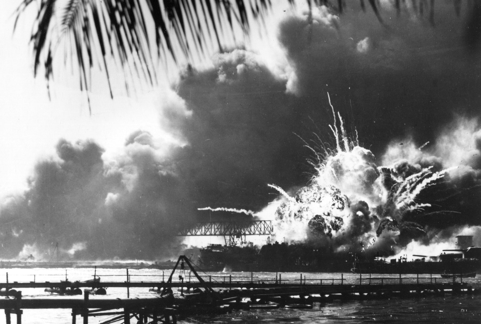 Black and white photograph framed by palm fronds: a huge fireball with streaks of burning debris bursts from a destroyer in a floating dry dock across the harbor, under towering clouds of black smoke, with a dredge line in the foreground.