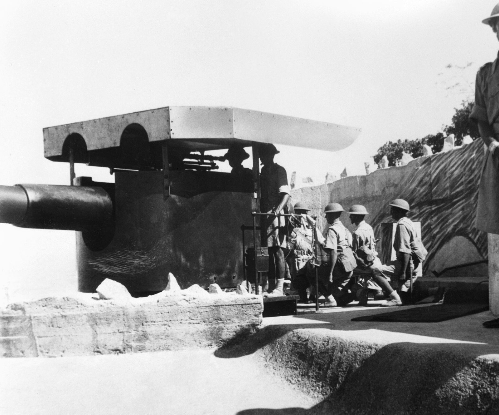 Black and white photograph of Indian gunners in steel helmets and shorts working a large coastal gun under a flat steel shield in a stone emplacement, with a British officer standing at the right edge of the picture.