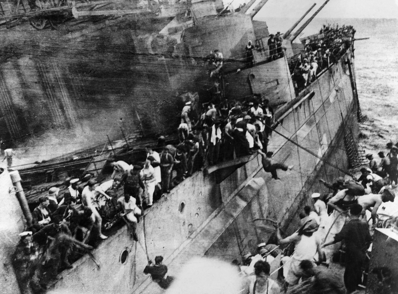 Black and white photograph looking down on the steeply listing side of a battleship as crowds of sailors, many in white, climb down ropes and gangways onto the deck of a destroyer alongside, one man swinging over the gap.