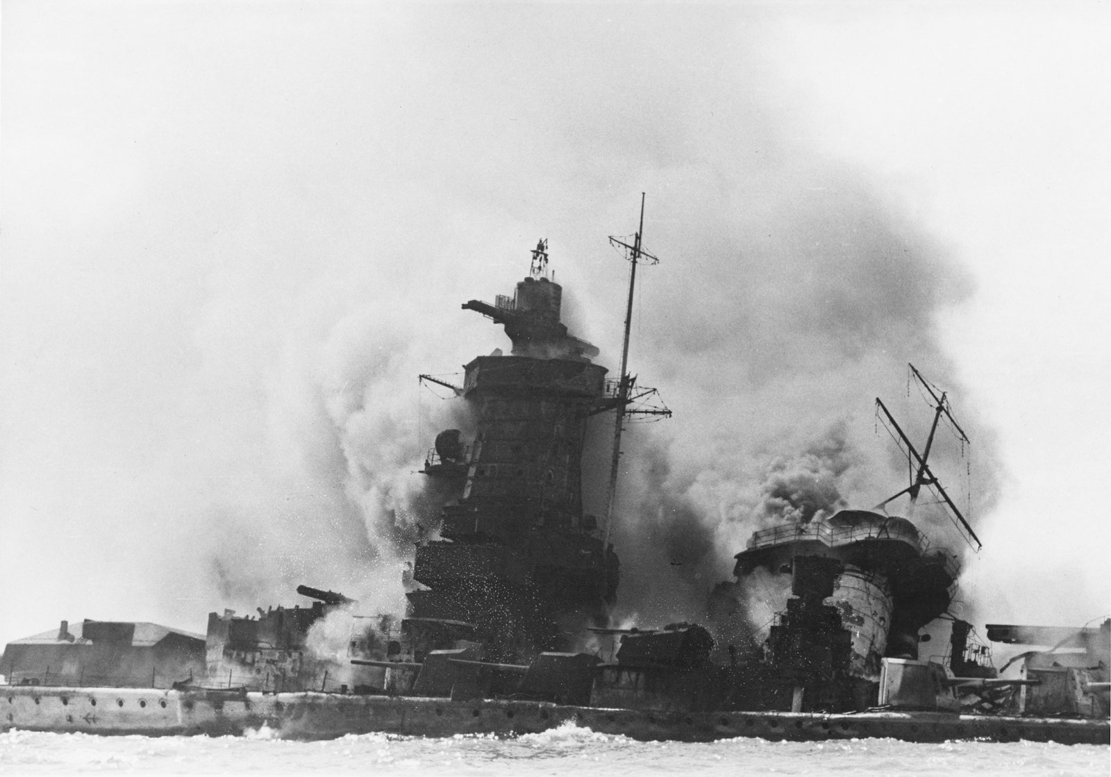 Black and white photograph of a wrecked warship low in the water, its tall tower superstructure, mast and guns wreathed in white and black smoke from fires and explosions.