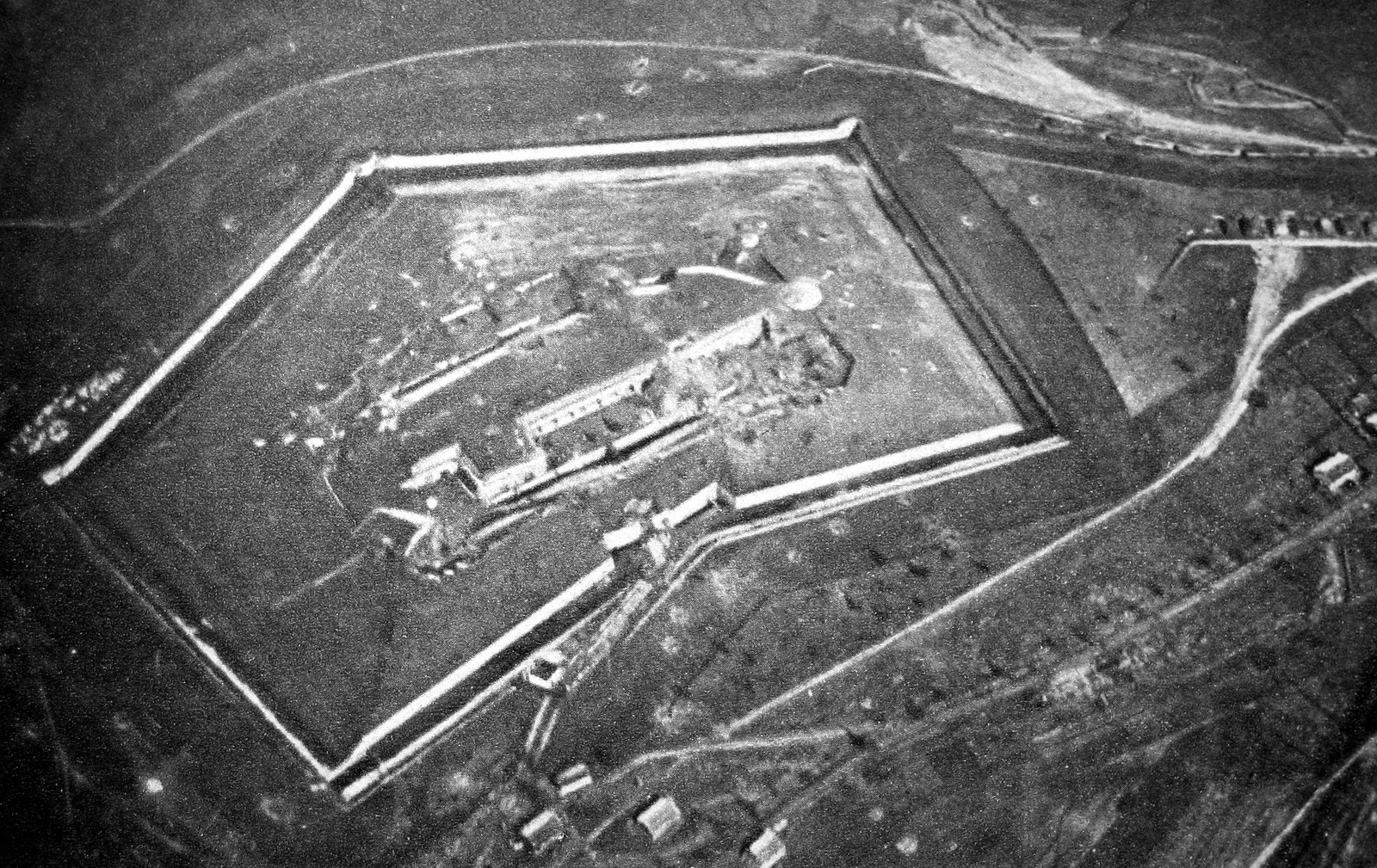 Black and white aerial photograph of a five-sided fort seen from above, its low barracks block inside a ditch with pale walls, surrounded by open ground marked with tracks and scattered shell holes.
