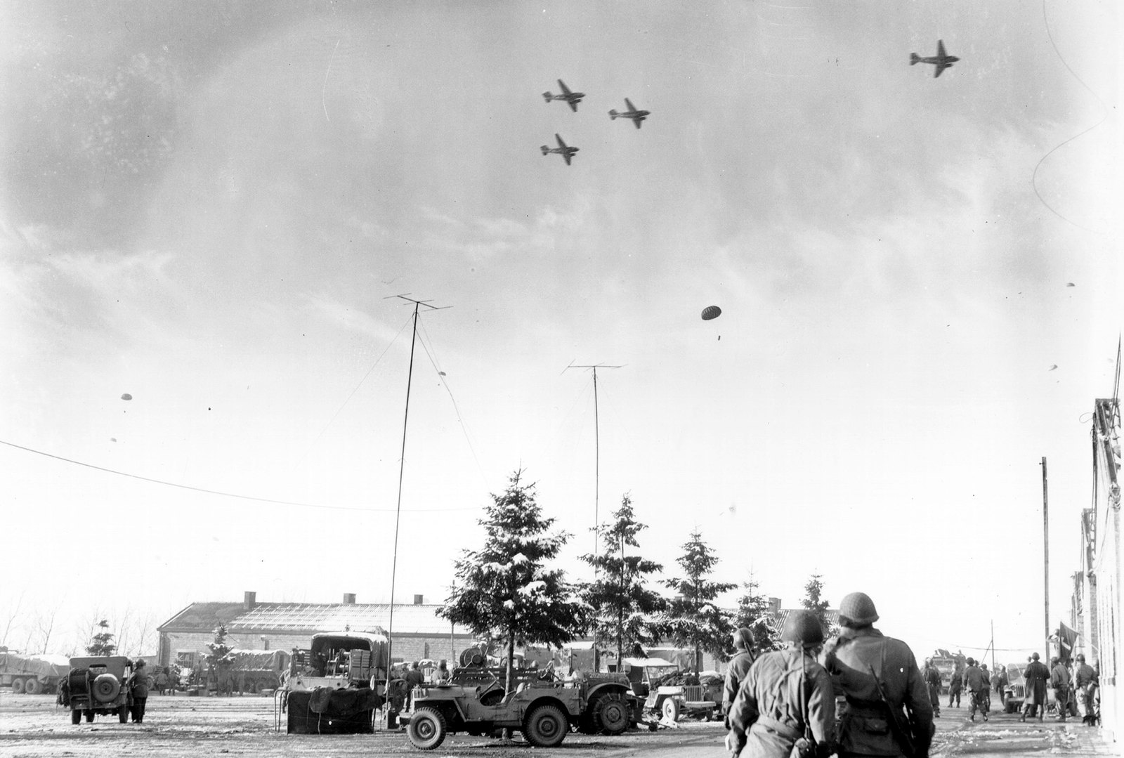 Black and white photograph: two American soldiers in helmets, seen from behind, watch twin-engine transport planes fly over a snowy square with parked jeeps and trucks, fir trees and low barracks, while a parachute drifts down.