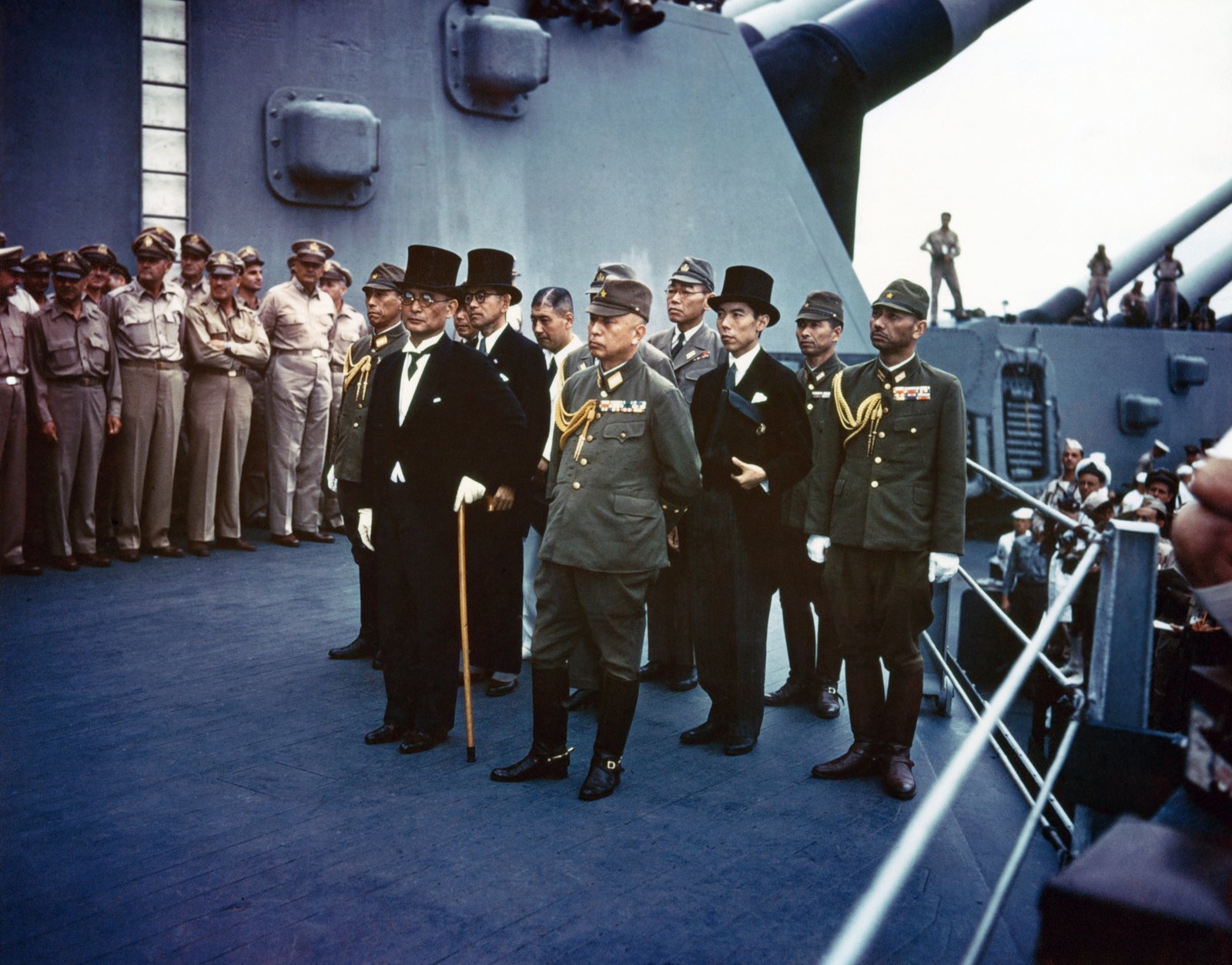Color photograph: the Japanese delegation, diplomats in top hats and morning coats and officers in uniform, stands in rows on the deck of USS Missouri, watched by American sailors and officers.