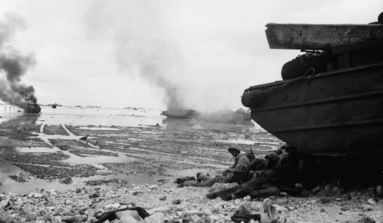 Marines take shelter under an amphibious DUKW on the beach at Peleliu in September 1944 while amphibious tractors burn on the reef behind them