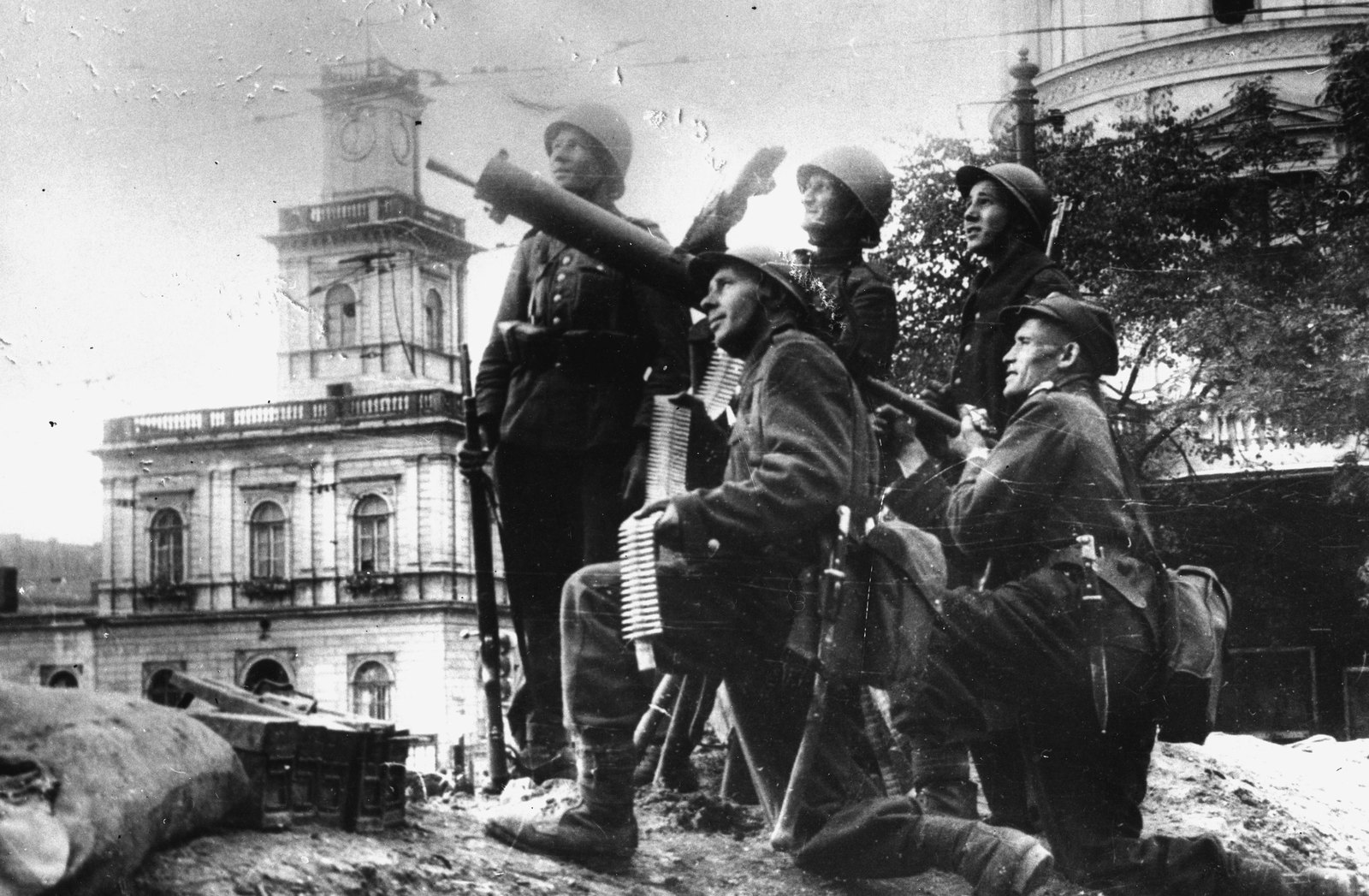 Czarno-biała fotografia pięciu polskich żołnierzy w hełmach na stanowisku z worków z piaskiem; jeden celuje z karabinu maszynowego w niebo, drugi trzyma taśmę z amunicją, w tle klasycystyczny budynek z wieżą zegarową.