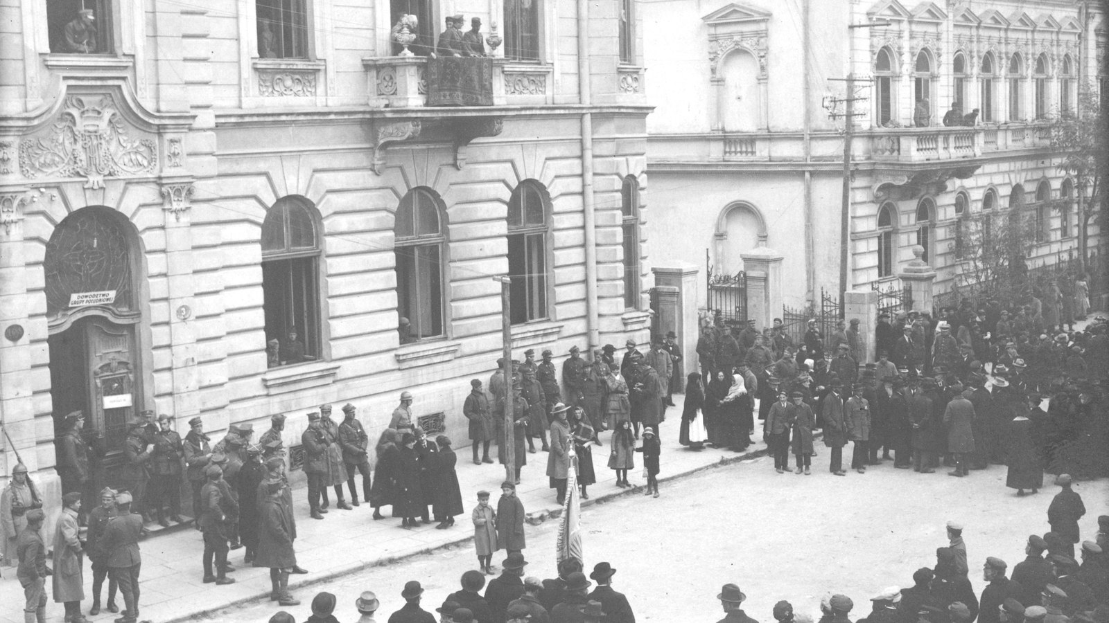 Czarno-białe zdjęcie ozdobnego kamiennego gmachu z tablicą nad łukowym wejściem; wzdłuż ściany stoją żołnierze w czapkach i stalowym hełmie, w oknach i na balkonie gapie, a ulicę przed budynkiem wypełnia tłum cywilów w kapeluszach i chustach, pośrodku zwinięty sztandar