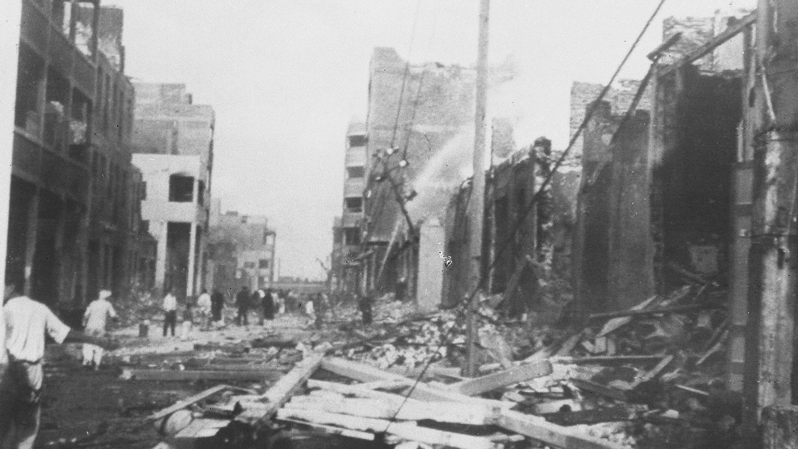 Black and white photograph of a wrecked street in Port Said, with broken beams and rubble in the foreground, burned-out buildings on both sides, a jet of water on a smoking ruin and people walking in the distance