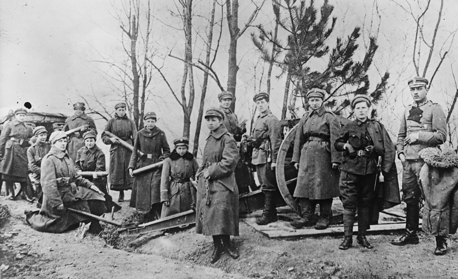 Czarno-biała fotografia grupy młodych polskich ochotniczek w płaszczach i czapkach z daszkiem, stojących przy dziale polowym wśród bezlistnych drzew, z oficerem po prawej.
