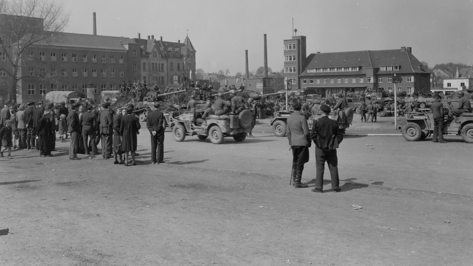 Black and white photograph of townspeople in coats watching as U.S. jeeps tow away surrendered German anti-tank guns on a square in Iserlohn, with brick buildings and factory chimneys behind, 16 April 1945.