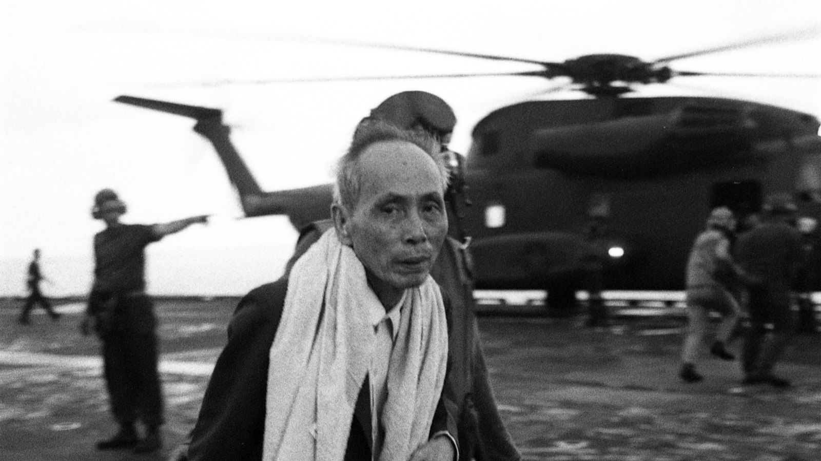 Black and white photograph of an elderly Vietnamese man with a towel round his neck and a smiling little girl walking across the flight deck of USS Hancock on 29 April 1975, with a CH-53 helicopter and Marine deck crew behind them.