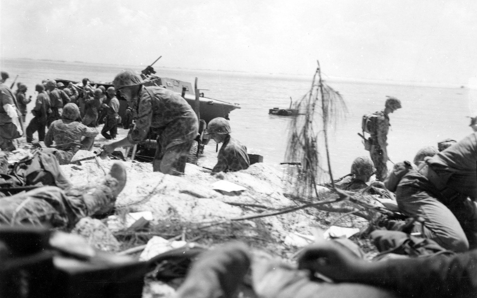 Black and white photograph of US Marines in camouflage on a sandy beach under fire, one reaching down in the foreground while others crouch and wade beside an amphibian tractor at the water's edge, the lagoon and reef behind