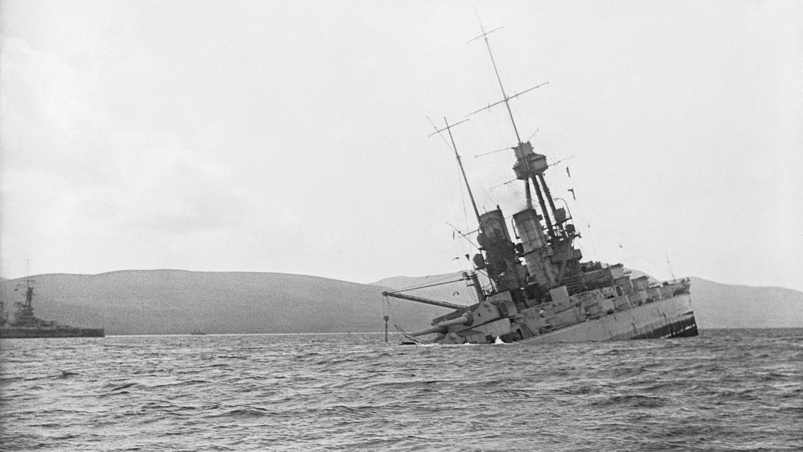 Black and white photograph of a German battleship listing heavily and sinking by the stern in choppy water, with hills and another warship on the horizon.