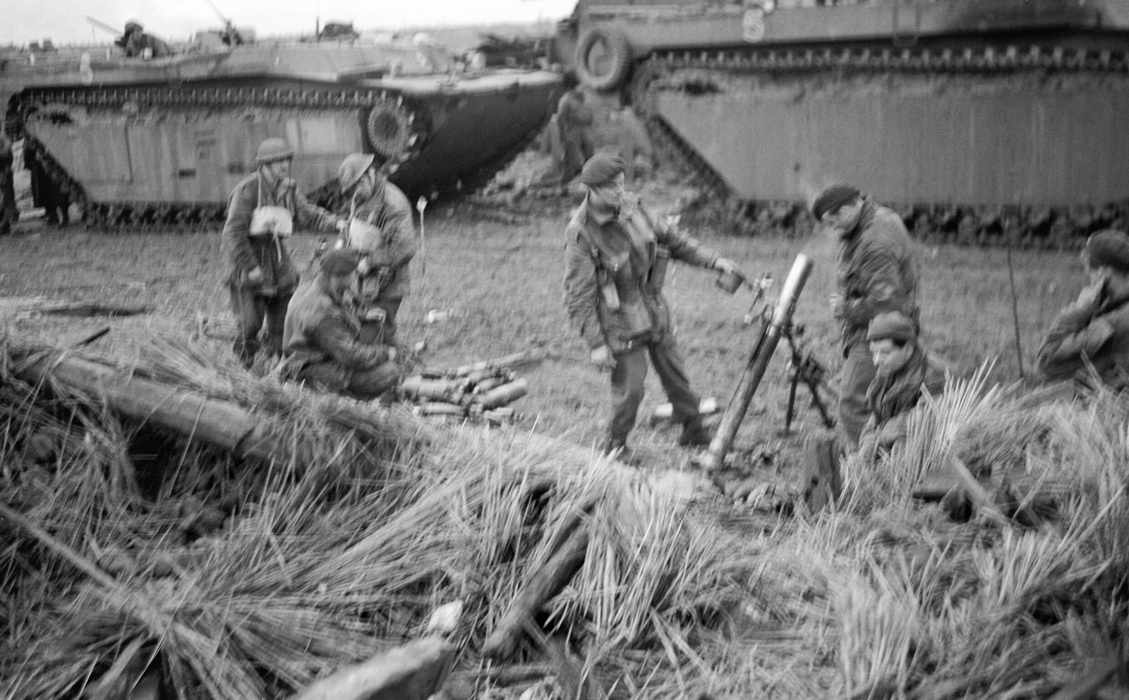 Black and white photograph of British soldiers in helmets and berets firing a mortar from a muddy, reed-strewn position, with two large amphibious tracked vehicles parked behind them.