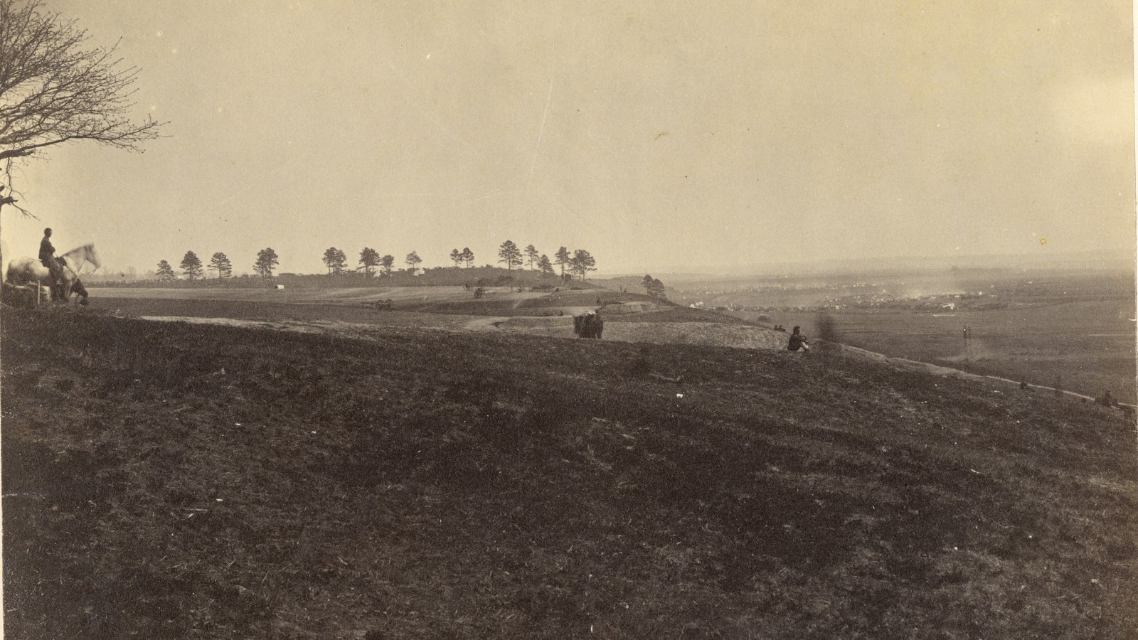 Faded 1863 photograph looking across the bare slope of Marye's Heights at Fredericksburg, with a rider and a few figures on the ridge, a line of pine trees on the horizon and the town and river plain in the distance.