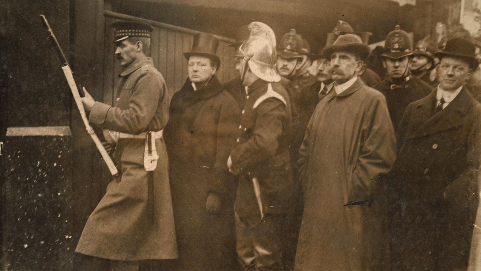 Old press photograph of a soldier aiming a rifle around a corner while Winston Churchill in a top hat, a fireman in a brass helmet, police officers and men in bowler hats stand behind him.