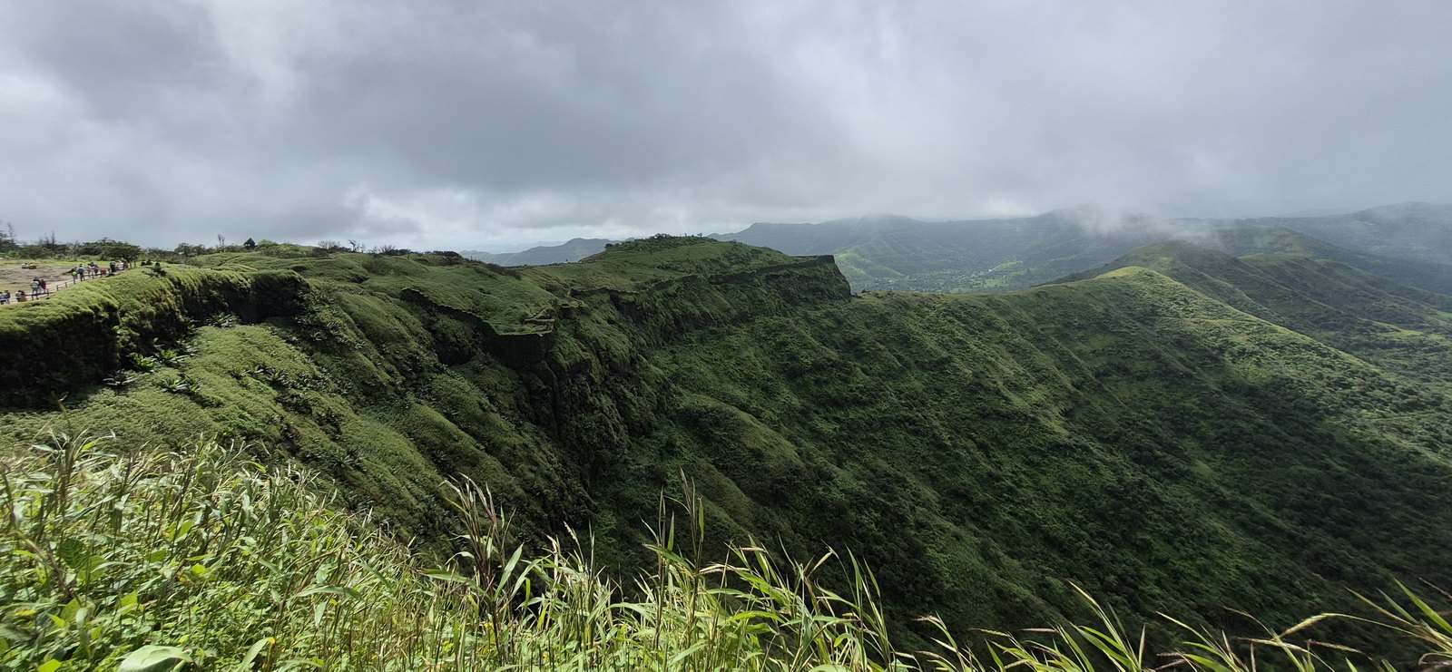 Green cliff-edged ridge of Sinhagad under heavy monsoon clouds, with visitors walking along the summit path on the left