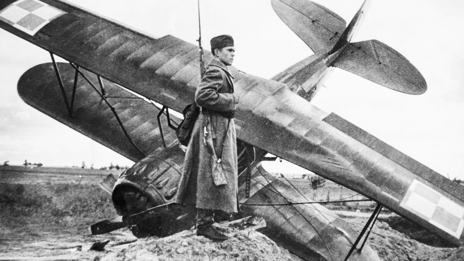 Black and white photograph of a Soviet soldier in a long greatcoat and fur cap, rifle with bayonet on his shoulder, standing guard beside a crashed biplane with the Polish checkerboard marking on its wings