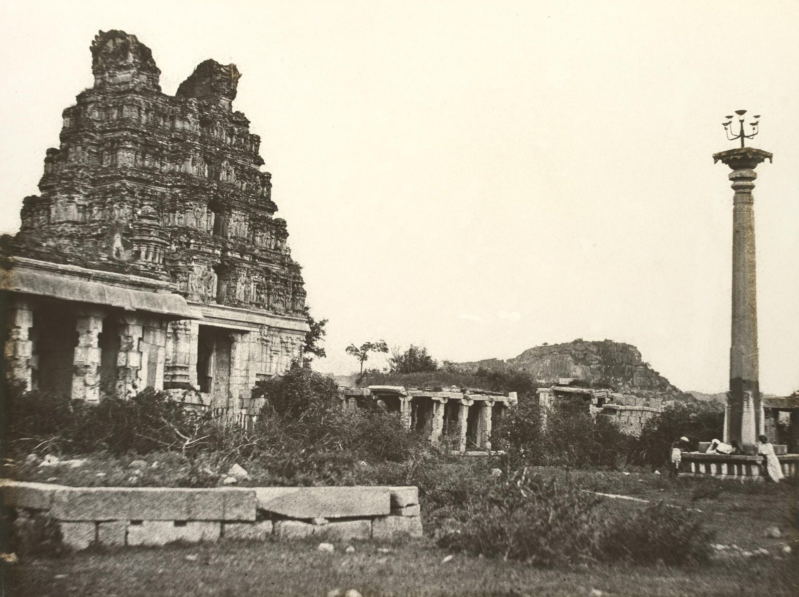 Ruined temple gateway tower beside a pillared hall, with a tall stone lamp column and scrub in the foreground, in an old sepia photograph
