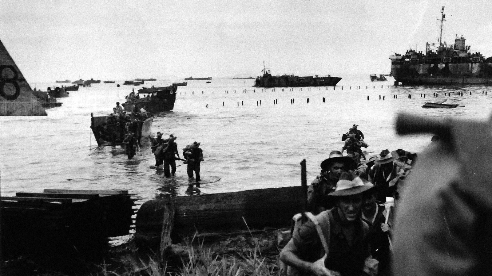 Black and white photograph of Australian soldiers in slouch hats wading ashore from landing craft at Tarakan on 1 May 1945, with rows of beach obstacles in the water and a large landing ship offshore.