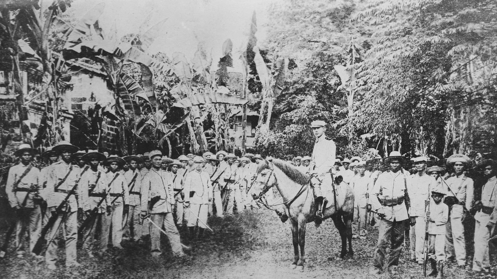 Old black and white photograph of a young officer in a white uniform on a horse in a village street, with two ranks of Filipino soldiers in hats and white uniforms holding rifles on either side and banana trees behind.