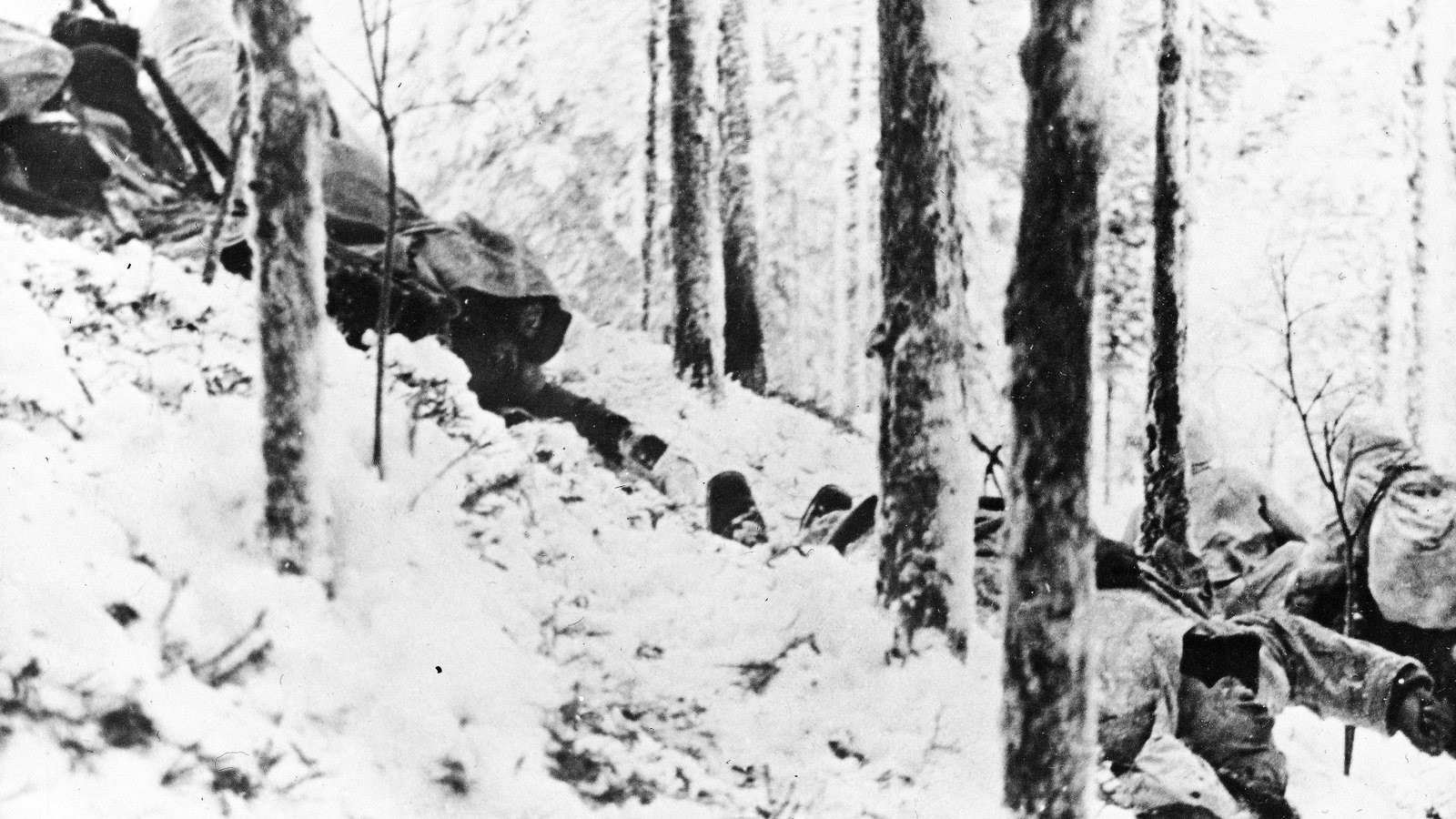 Black and white photograph of Finnish soldiers in white snow capes lying among snow-covered pines on a slope, rifles ready, during the attack at Tolvajärvi.