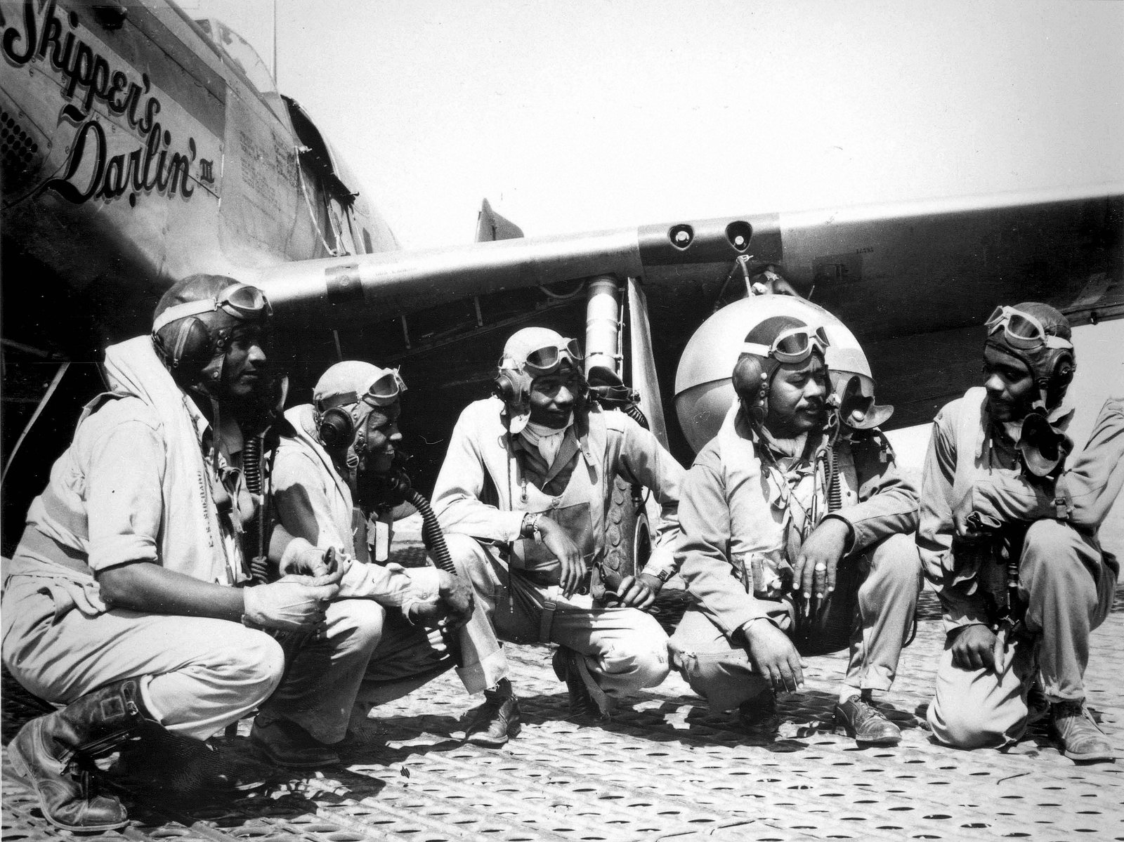 Five pilots of the 332nd Fighter Group in flying helmets crouch beneath the wing of a P-51 Mustang at Ramitelli, Italy, in 1944
