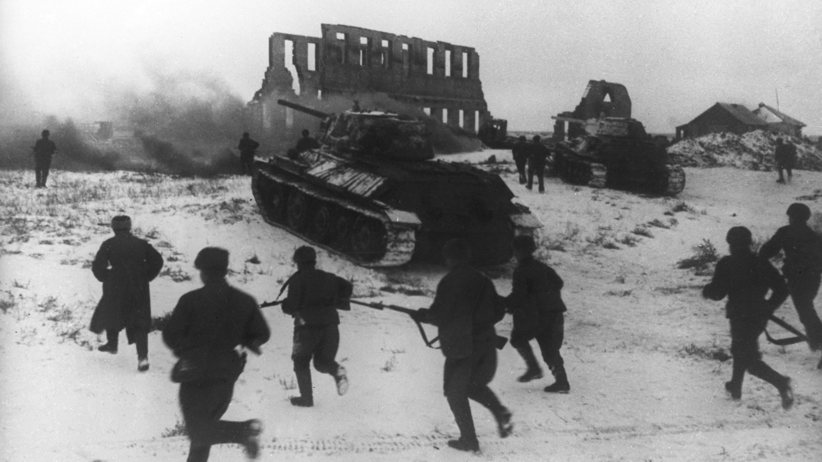 Black and white wartime photograph of Soviet infantry running across a snowy field behind two T-34 tanks, with a ruined building and smoke in the background