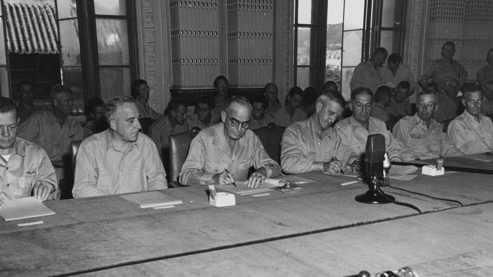 Black and white photograph of American admirals and generals seated in a row at a long table in a high-windowed hall, one of them signing a document, with a microphone in front of them and officers standing behind.