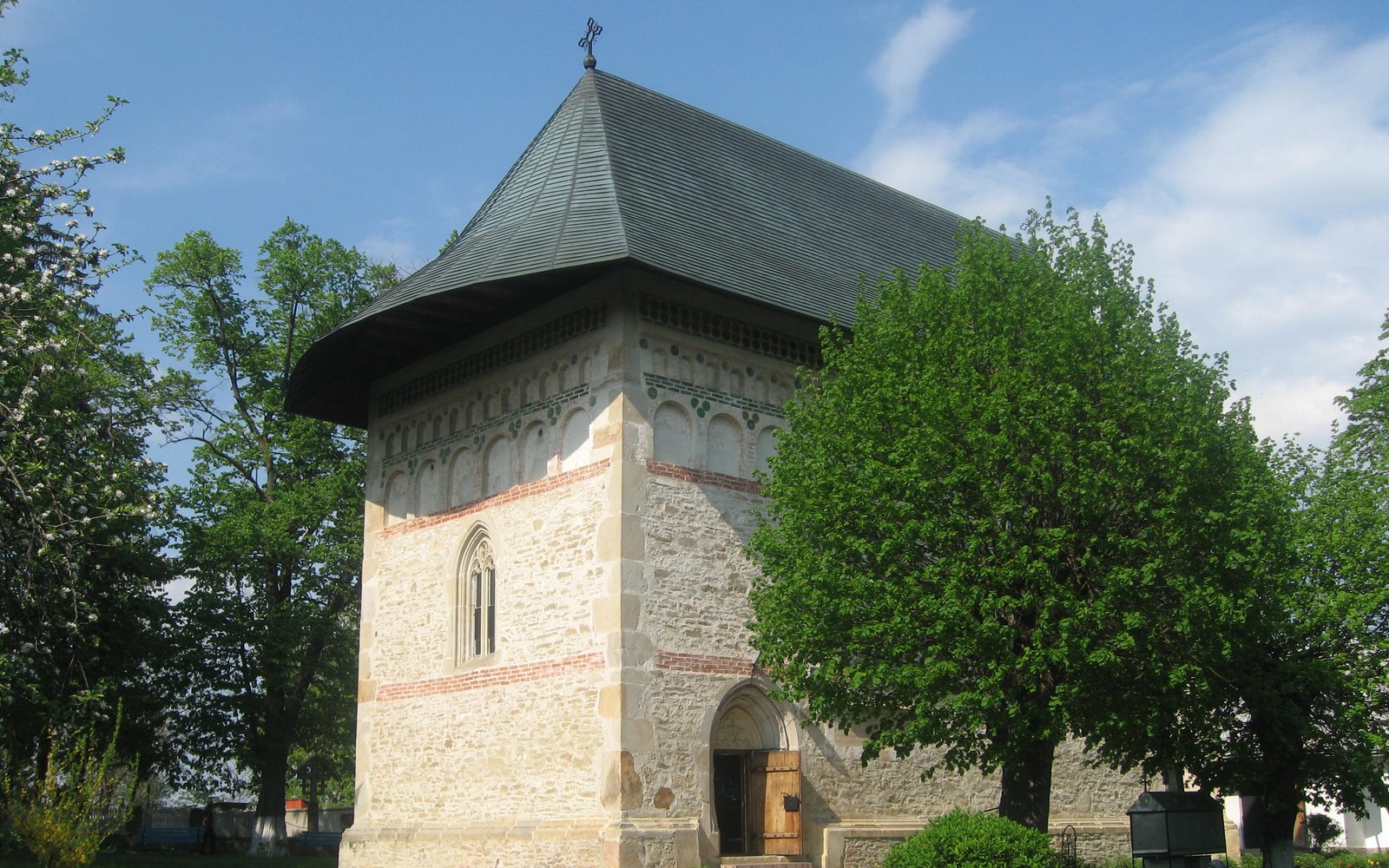 Photograph of a small medieval stone church with a steep dark shingled roof and decorative brick bands, surrounded by green trees under a blue sky.