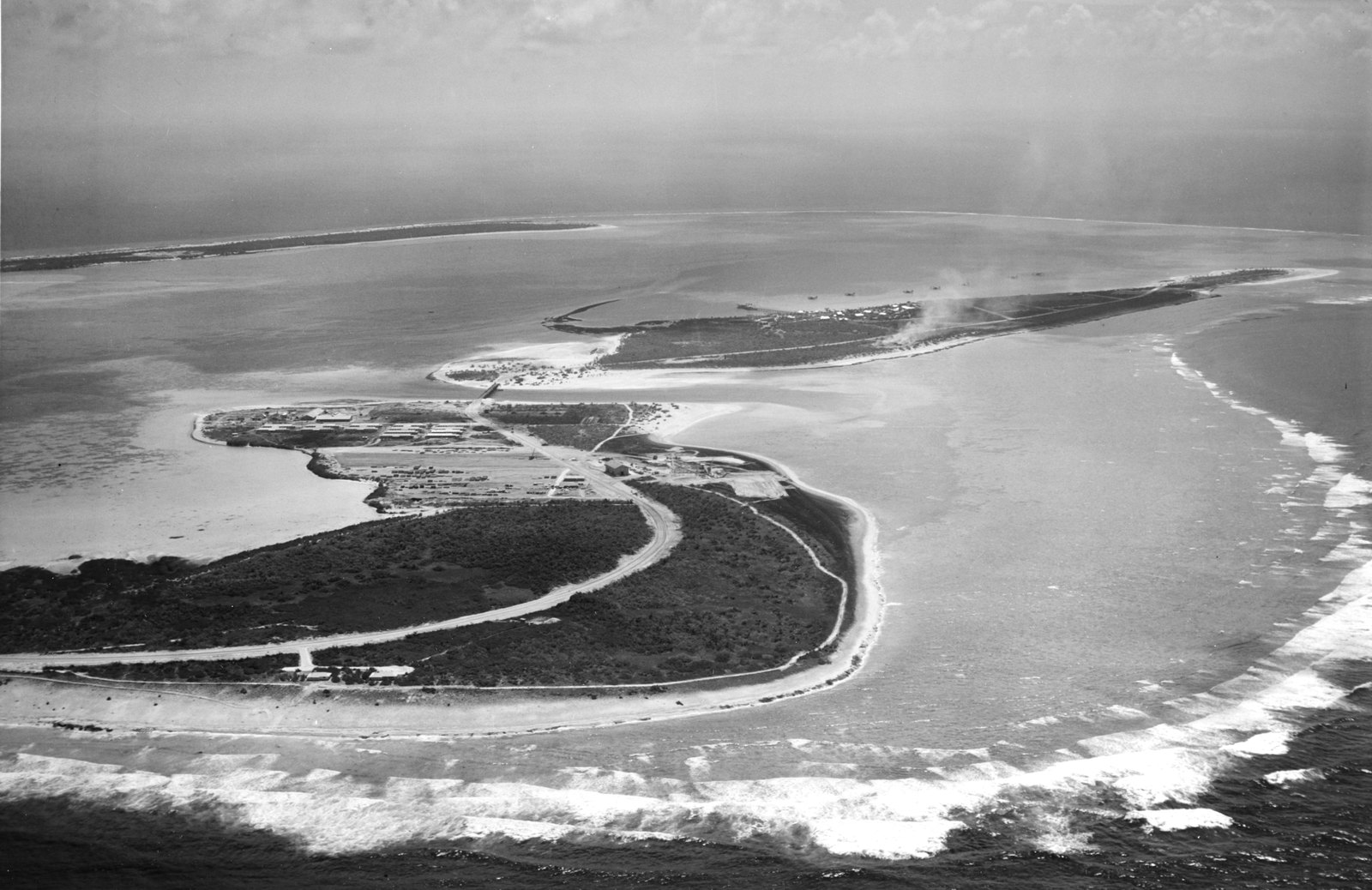 Black and white oblique aerial photograph of a curving wooded islet with a road, rows of camp buildings and a bridge to a long narrow second islet, a third low islet on the horizon and white surf breaking along the reef