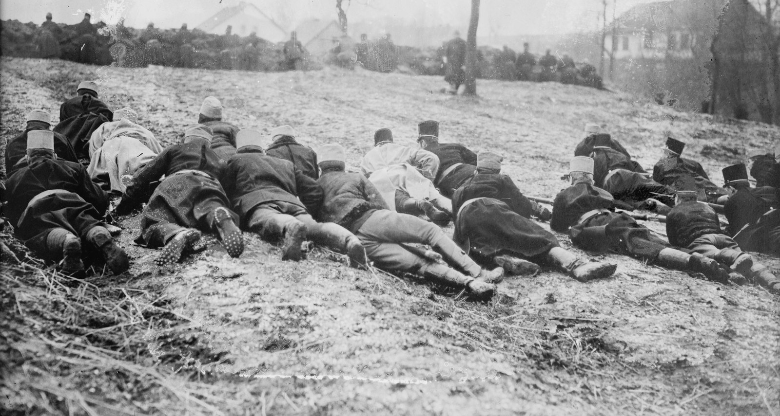 Black and white photograph: a line of soldiers in dark greatcoats and soft caps lie prone on a frosty slope, rifles forward, with more troops, bare trees and a farmhouse behind them.