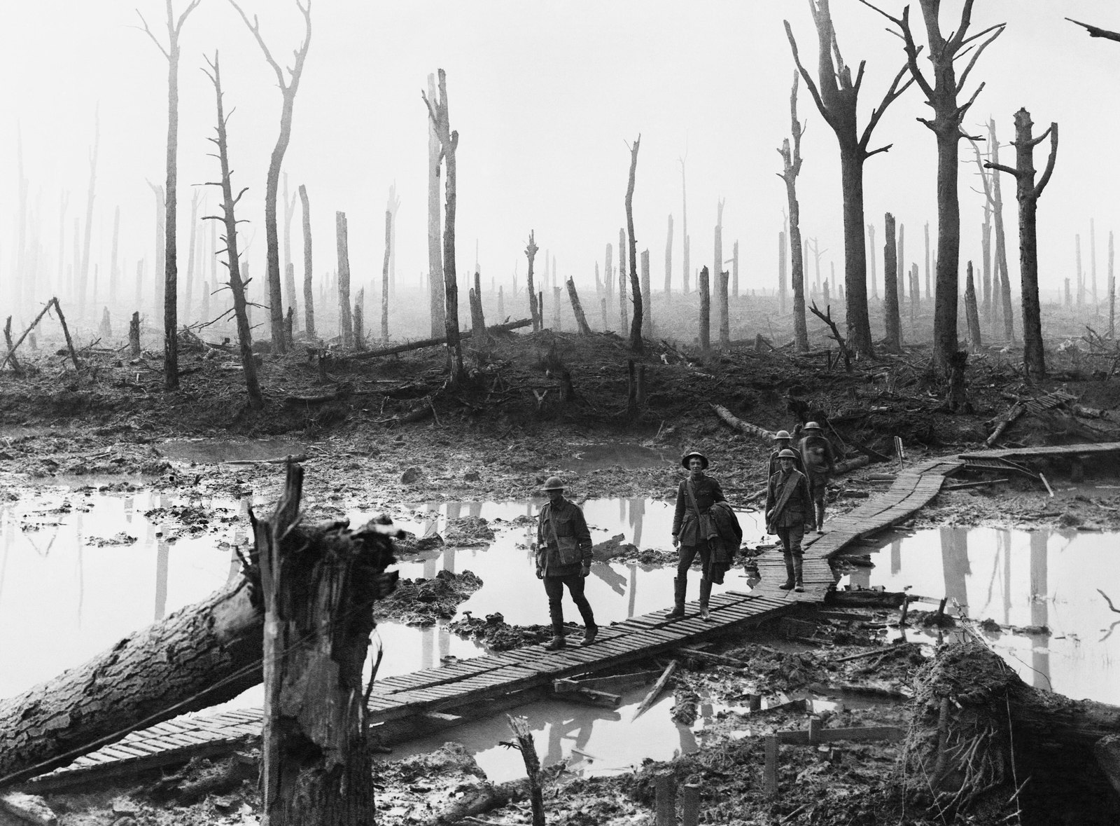 Black and white photograph: five soldiers in steel helmets walk along a duckboard track through a flooded, shell-torn wood of bare, shattered tree trunks in the mist.