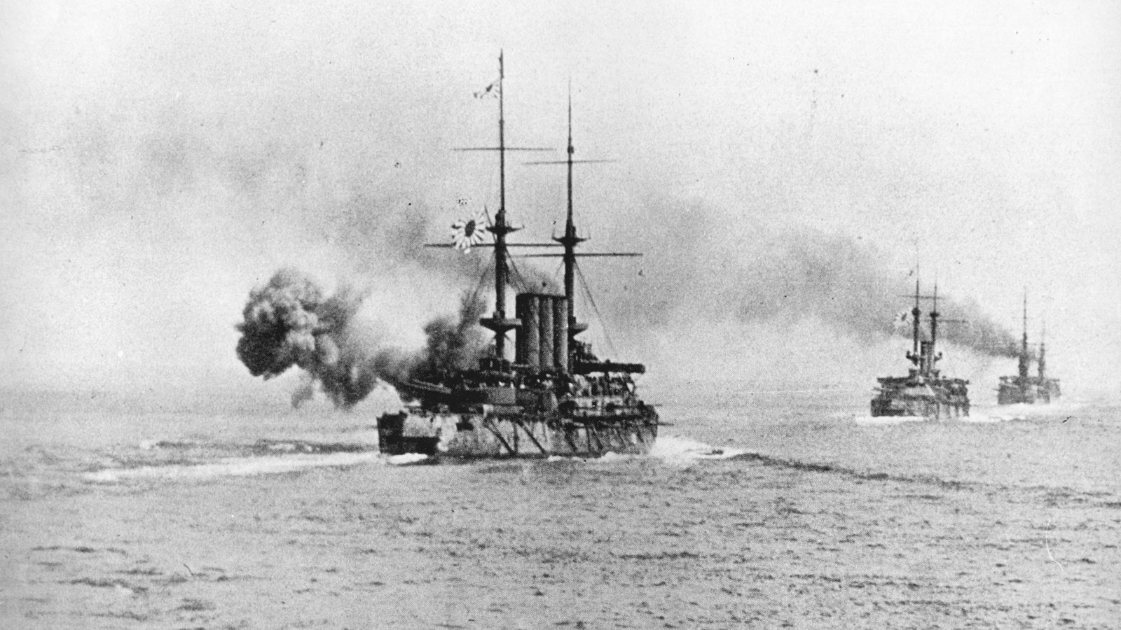 Black and white photograph from the Battle of the Yellow Sea: the Japanese battleship Shikishima fires a gun in a cloud of smoke as two more battleships follow in line across a hazy sea.