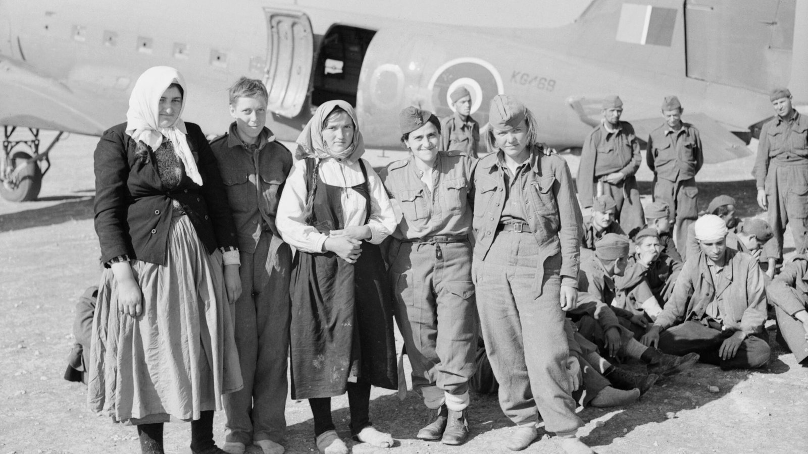 Black and white photograph of partisan and civilian women and young soldiers standing in front of a twin-engine transport aircraft, with wounded men sitting on the ground.