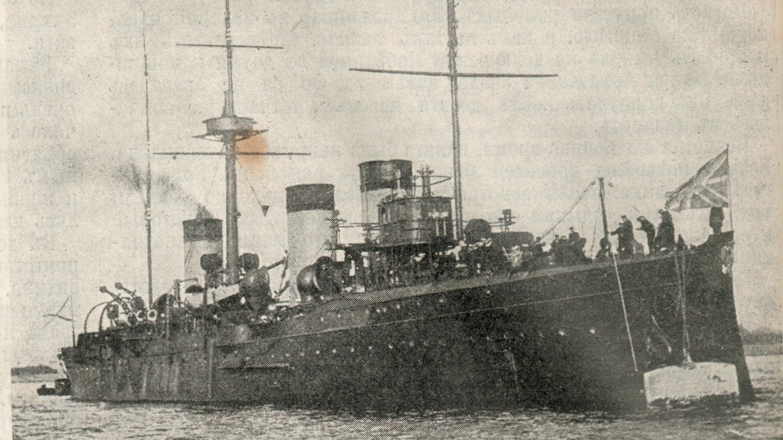Old halftone photograph of a dark three-funneled warship with tall masts lying at anchor, sailors on the forecastle and a flag flying at the bow