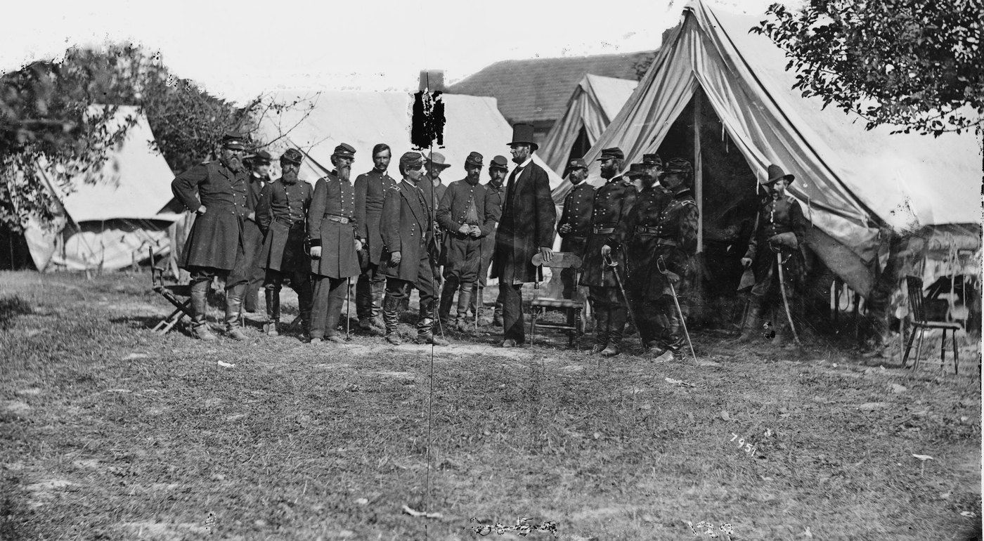 Black and white photograph: Abraham Lincoln in a dark suit and top hat stands among a row of uniformed Union officers in front of army tents.