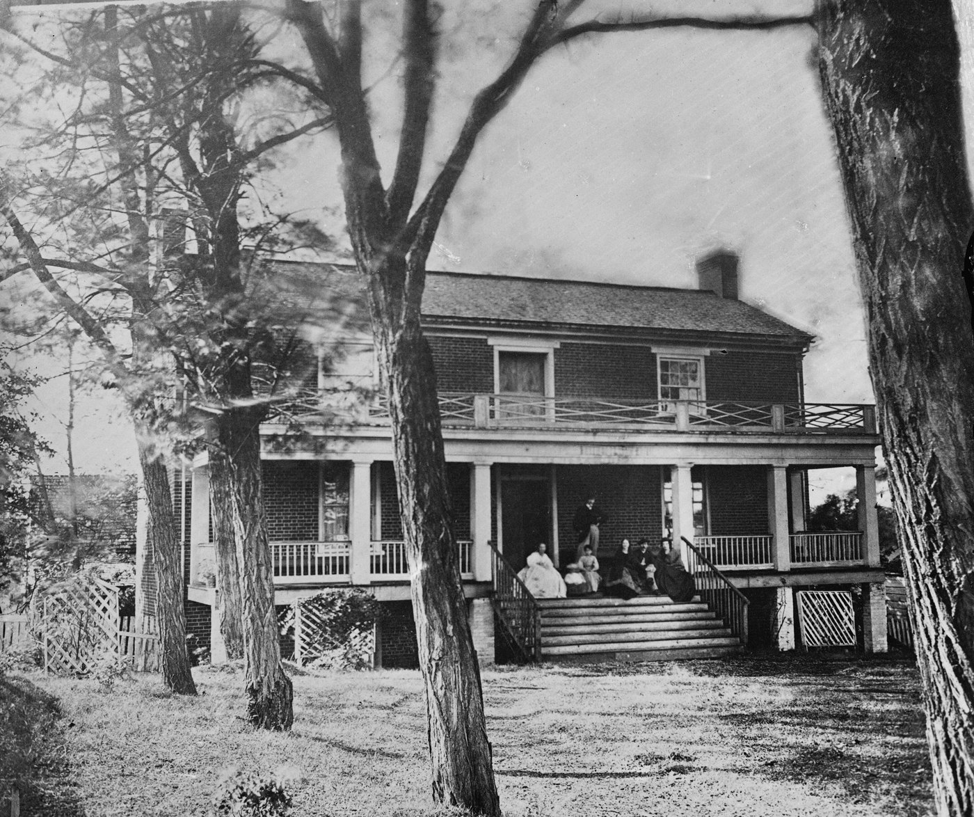 Black and white photograph of a two-story brick house with a long double porch and wooden steps, seen between tall trees, with several people seated on the steps.