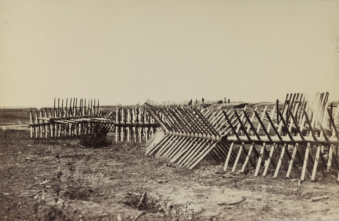 Sepia photograph of rows of sharpened wooden stakes set in crossed frames in front of earthworks, with a few soldiers standing on the parapet in the distance.