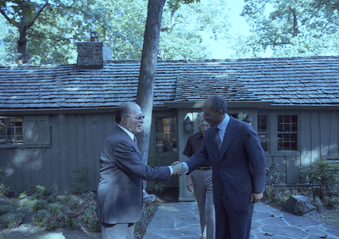 Color photograph of two men in suits shaking hands on a stone path in front of a wooden cabin under tall trees: on the left an older man with glasses, on the right a taller man, with a third man in a brown shirt standing between them.