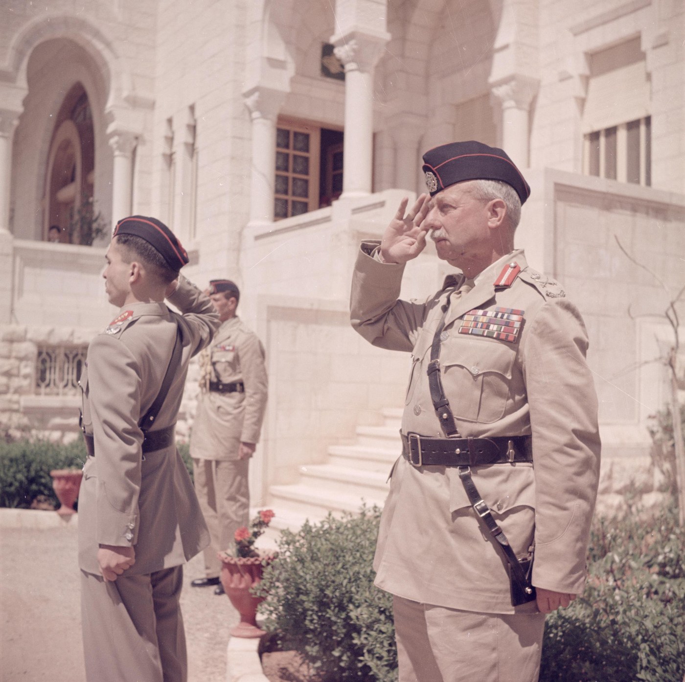 Color photograph of two officers in khaki uniforms and dark berets saluting side by side in front of a pale stone palace with arches: on the right an older officer with a white mustache and rows of medal ribbons, on the left a young man in profile, with a third officer behind them.