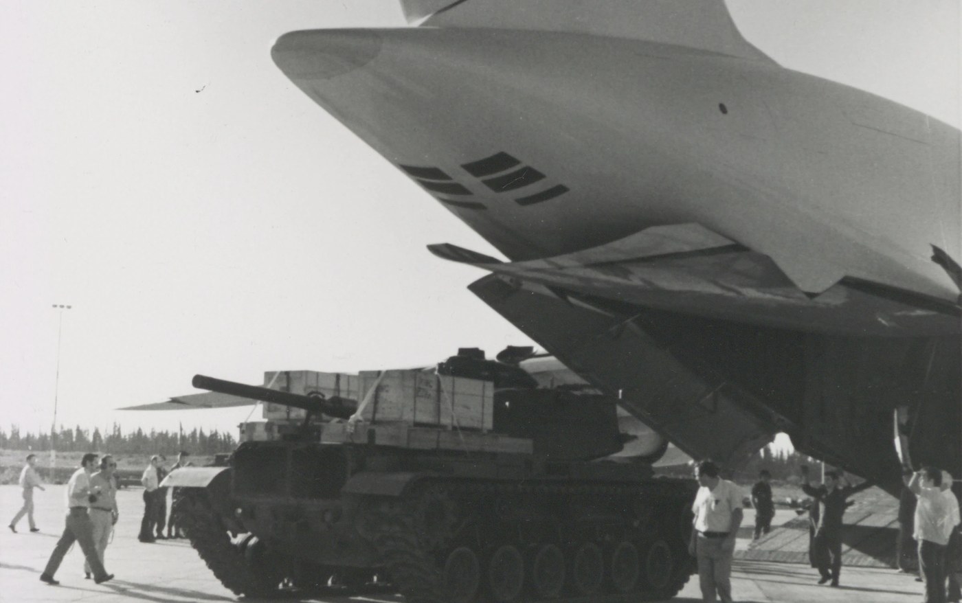 Black and white photograph of an M60 tank, its hull stacked with wooden crates, rolling out from under the raised tail of a huge US Air Force C-5 transport plane on an airfield apron, while men in shirtsleeves walk alongside and guide it.