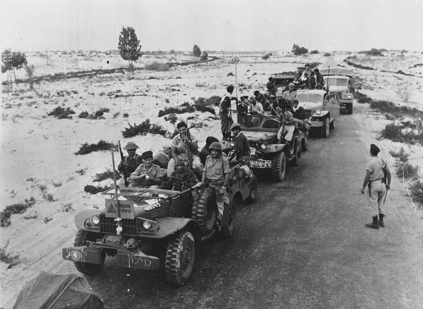 Black and white photograph of a column of open military jeeps and trucks full of soldiers with rifles on a straight desert road through scrubby sand, with a soldier in a beret standing at the roadside on the right.